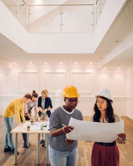 Two female architect looking at blueprint at architecture studio. Portrait of african american and vietnamese woman engineer in office with their colleagues having a meeting in the background.
